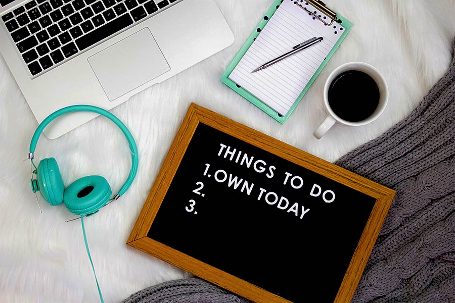 letrero con el mensaje "cosas por hacer: 1. adueñar el día" sobre una alfombra al lado de una agenda, audífonos, café y una laptop