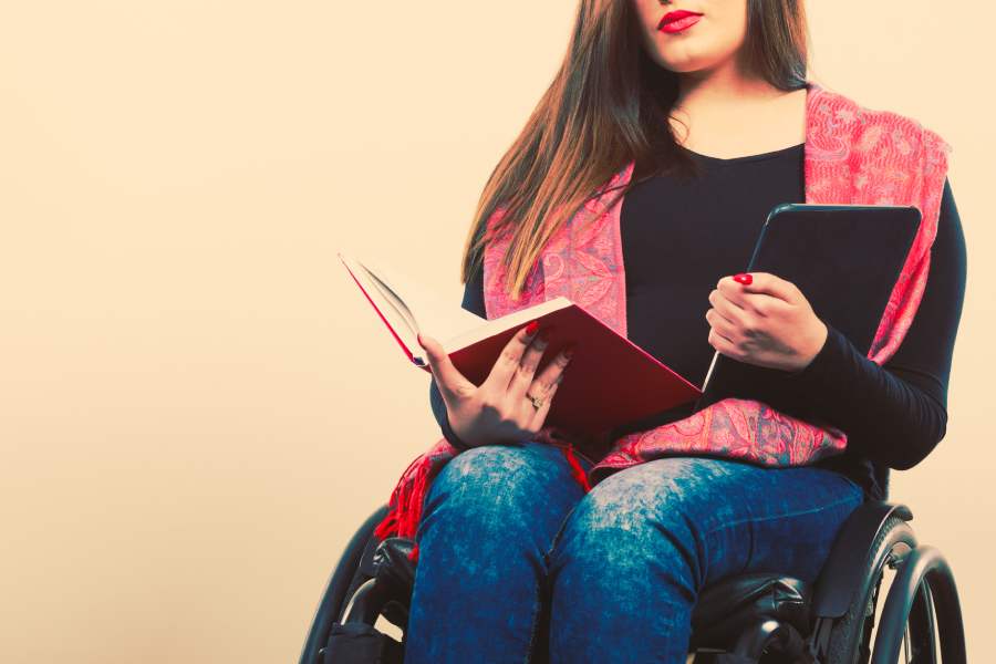 Joven mujer en silla de ruedas, sosteniendo en una de sus manos un libro impreso, y en la otra un lector de libros digitales