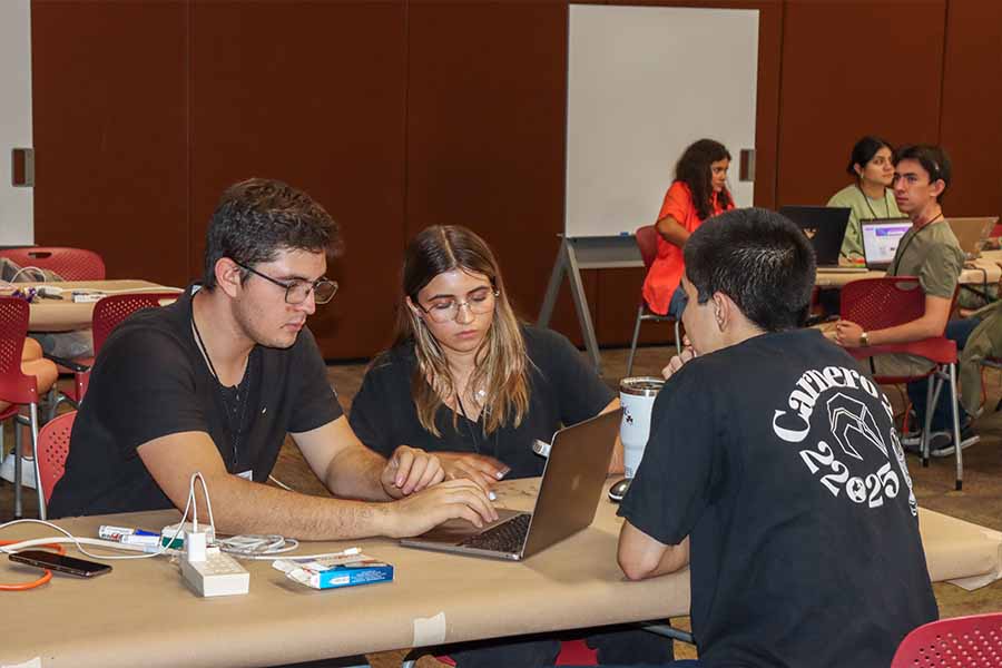 Tres alumnos sentados en una mesa con computadoras trabajando.