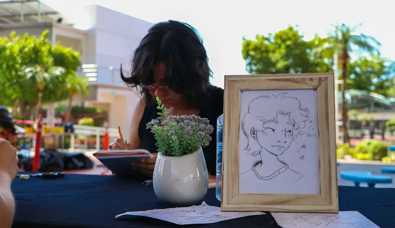 Chica dibujando sobre una mesa y enfrente de ella se ve un dibujo de una cara en un marco de madera. 