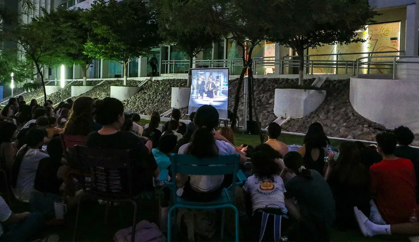 Grupo de estudiantes sentados en un jardín viendo una película proyectada sobre una pantalla blanca. 