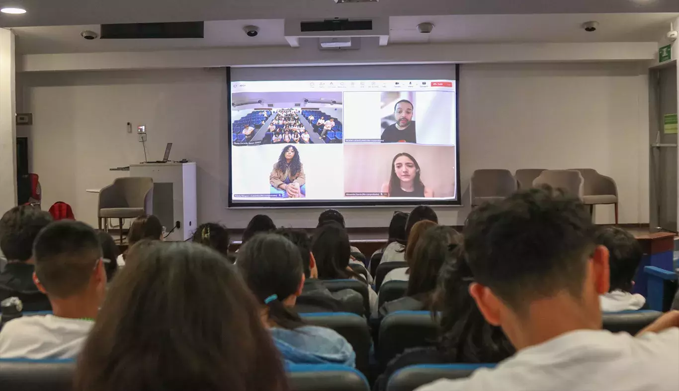 Público en auditorio volteando hacia una pantalla que muestra una videollamada en proceso.