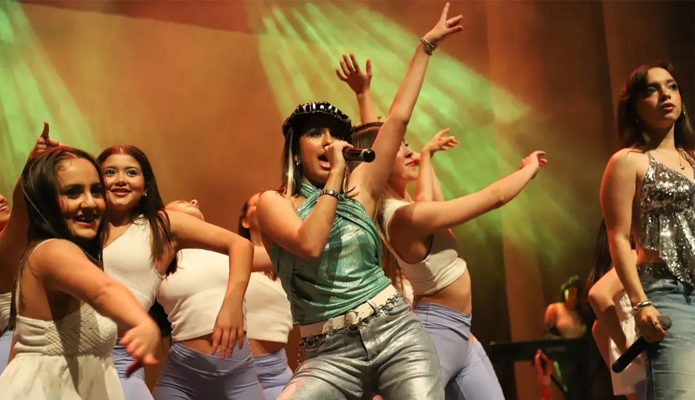 Chica con blusa brillante verde y boina canta rodeada de bailarinas vestidas de blanco.