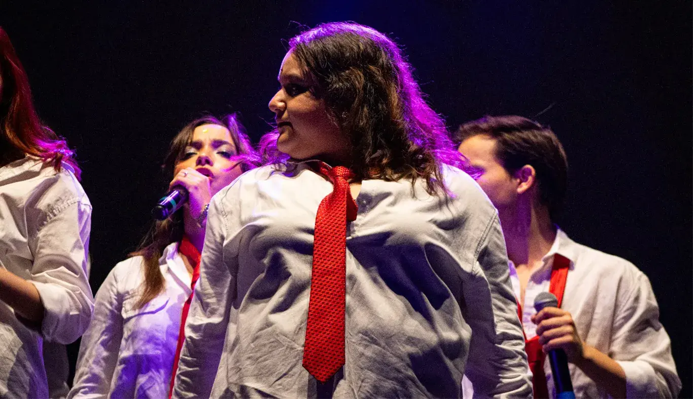 Chica vestida estilo colegial cantando en el escenario volteando hacia la izquerda.