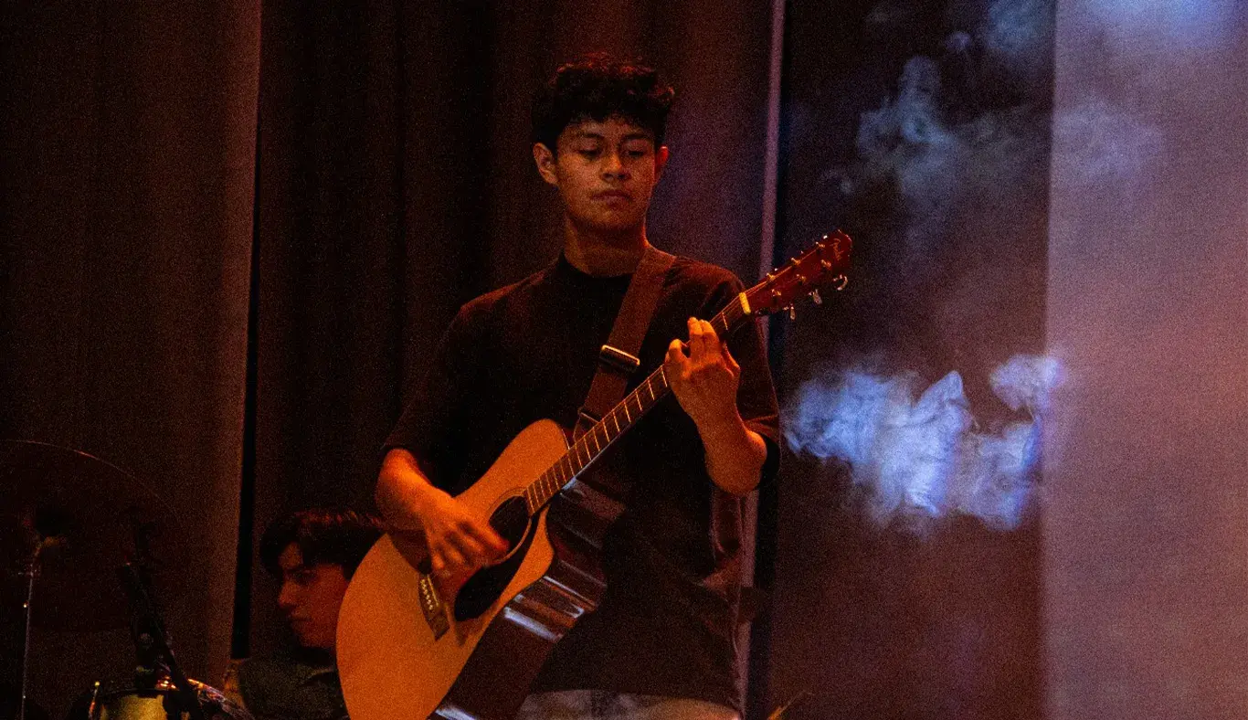 Chico tocando la guitarra acústica en escenario con humo alrededor. 