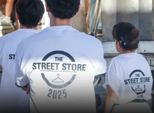 Vista de espaldas de tres voluntarios portando camisetas blancas con un logo que dice Street Store 2025