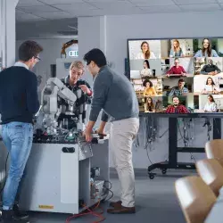 Laboratorio de clase de ingeniería, con 3 alumnos presenciales y diversos estudiantes en modalidad virtual que se visualizan en una gran pantalla al fondo, muestra del aprendizaje ciber-físico