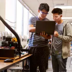 2 jóvenes estudiantes en un laboratorio; uno de ellos sostiene una laptop en su mano y señala su pantalla