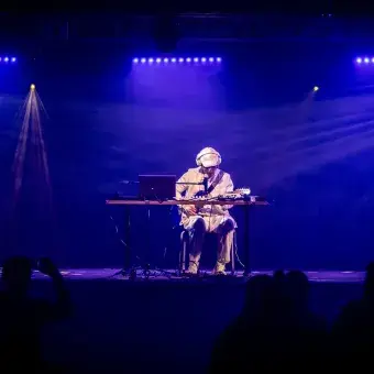 Jesús Alejandro, estudiante y músico, tocando en un escenario iluminado en tonos azules durante su presentación. Sentado con su equipo, la audiencia observando su interpretación.