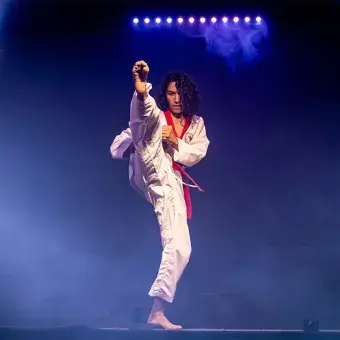 Rod Mendoza, estudiante y artista marcil, ejecutando una patada en su presentación, vestido con su uniforme de y rodeado de luces.